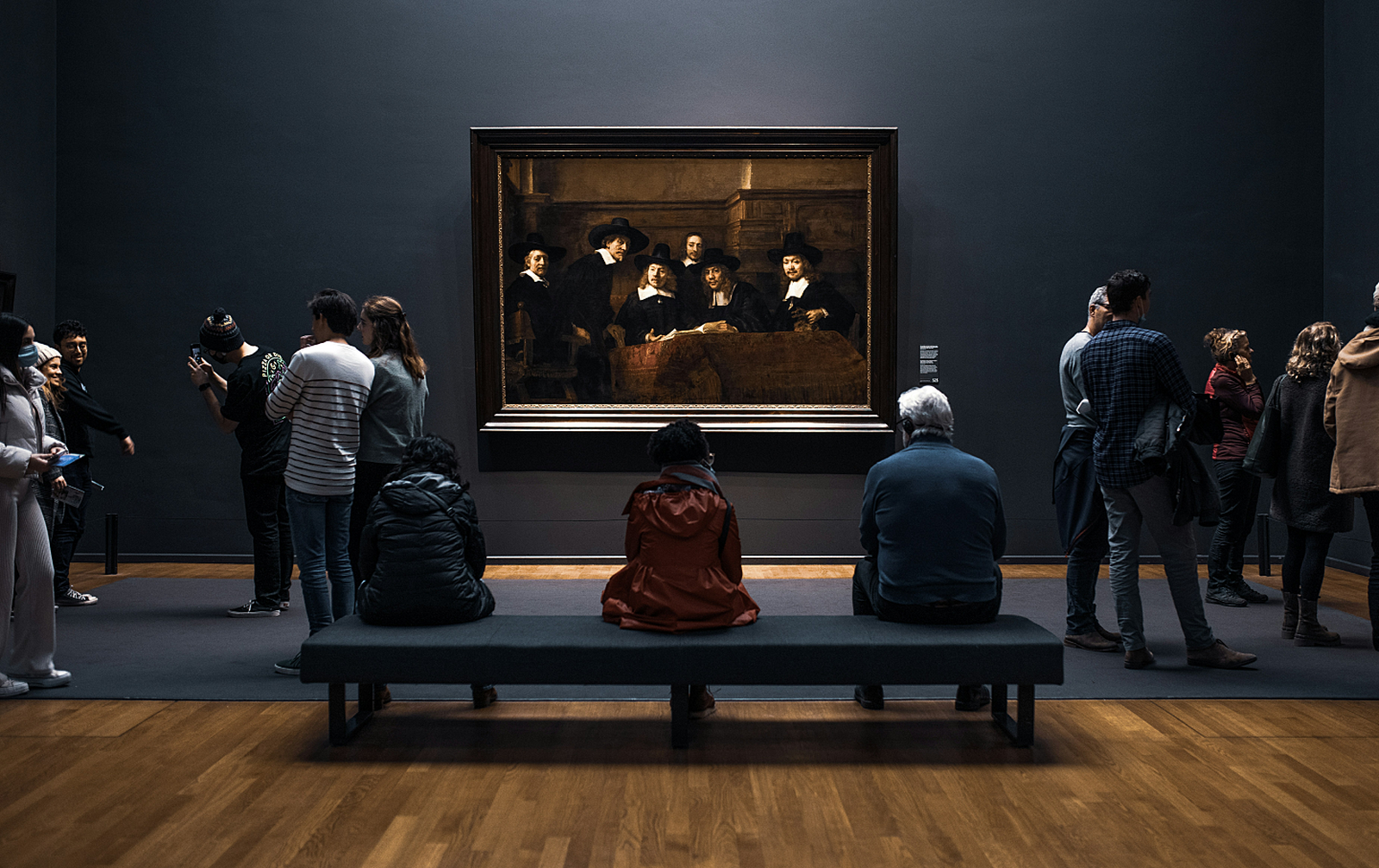 Image of 3 people on a bench with others crowded around, viewing an artwork, displayed in a Gallery.